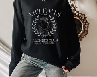 Peut inclure: Sweat-shirt noir avec le mot "ARTEMIS" au-dessus d'une couronne de laurier et le texte "ARCHERS CLUB GOLDEN BOW MEMBER." Le motif est gris clair. Le sweat-shirt est porté avec un jean bleu et une chaîne dorée.