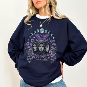 Crossroads Over Chaos sweatshirt featuring a moon goddess design, triple moon phase, violet flowers, butterfly, and the empowering phrase "Crossroads Over Chaos." Available in black, navy, maroon, and heather grey. Perfect for witchy aesthetic lovers and celestial symbolism fans.