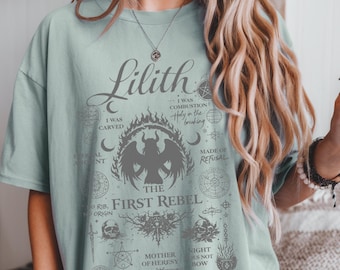 May include: A light green t-shirt with the word "Lilith" and various gothic-style illustrations. The design includes a central image of a winged figure, skulls, and text such as "The First Rebel" and "Mother of Heresy".