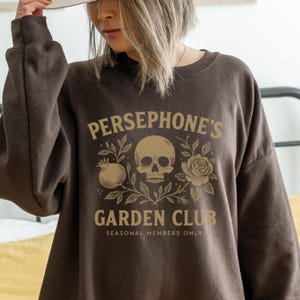 Sweatshirt featuring skull, pomegranate, and floral elements with text “Persephone’s Garden Club