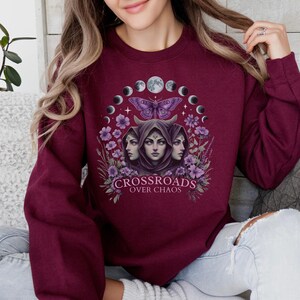 Crossroads Over Chaos sweatshirt featuring a moon goddess design, triple moon phase, violet flowers, butterfly, and the empowering phrase "Crossroads Over Chaos." Available in black, navy, maroon, and heather grey. Perfect for witchy aesthetic lovers and celestial symbolism fans.