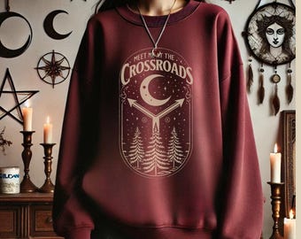 Może przedstawiać: Bordowa bluza z napisem "Meet Me at the Crossroads" i grafiką półksiężyca. Projekt zawiera strzałki i sosny. Bluza ma okrągły dekolt i długie rękawy.