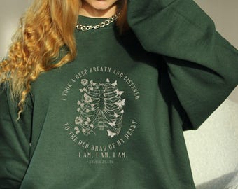 Può includere: Felpa verde scuro con la scritta "I TOOK A DEEP BREATH AND LISTENED TO THE OLD BRAG OF MY HEART. I AM. I AM. I AM." e un disegno di gabbia toracica con fiori e farfalle. La citazione è di Sylvia Plath.
