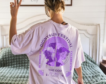 May include: A light purple t-shirt with a graphic design featuring the text "Dionysus Festival Celebration" and the words "The God of Wine" in a circular design. The design includes two female figures in purple.