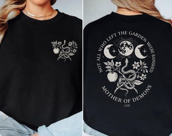 May include: Black sweatshirt with a small front graphic of an apple, snake, and flowers. The back features a circular design with moons, a snake, apple, and text that reads "NOT ALL WHO LEFT THE GARDEN WERE BANISHED MOTHER OF DEMONS".