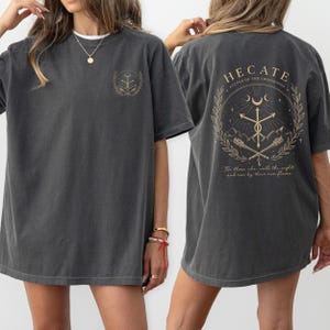 May include: Dark gray t-shirt with a round neck. The front features a gold emblem. The back displays the text "HECATE, KEEPER OF THE CROSSROADS" within a gold circular design, along with the phrase "For those who walk the night and rise by their own flame."