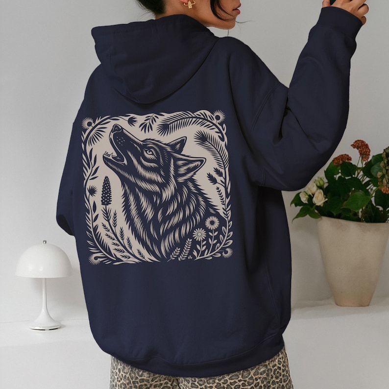 Model wearing a hoodie with a large linocut-style howling wolf illustration on the back, surrounded by botanical elements. Design shown in black, forest green, maroon, and dark chocolate color options.