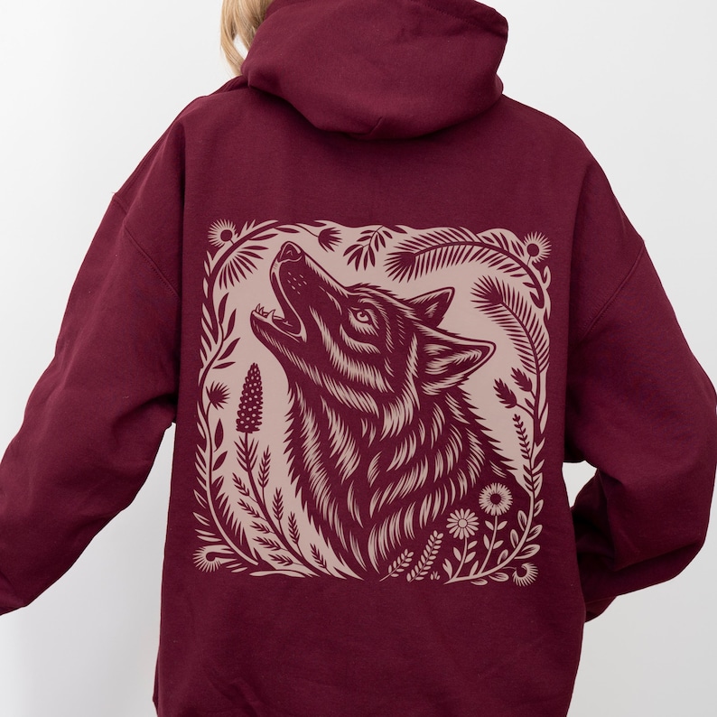 Model wearing a hoodie with a large linocut-style howling wolf illustration on the back, surrounded by botanical elements. Design shown in black, forest green, maroon, and dark chocolate color options.