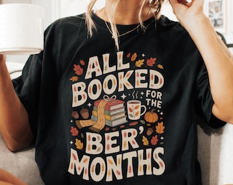 May include: Black t-shirt with the text "ALL BOOKED FOR THE BER' MONTHS" in a fall-themed design. The graphic includes books, a mug, a pumpkin, and autumn leaves in shades of orange, brown, and cream.