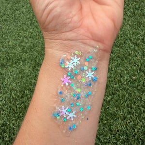 May include: A clear gel base containing a mixture of iridescent snowflake-shaped sequins, small blue star-shaped glitter, and tiny circular neon orange and green confetti pieces applied in a patch on the skin.
