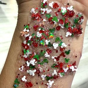 May include: A clear gel base containing a festive mix of red, green, and white glitter. The glitter includes small, distinct shapes resembling mouse heads, along with fine, shimmering particles that create a reflective, multi-colored effect.