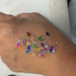 May include: A clear gel base containing a mixture of fine silver glitter and larger, multi-colored, iridescent sequins shaped like mouse heads in shades of red, blue, green, yellow, and purple, applied in a cluster on the skin.