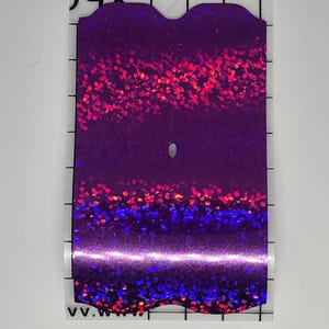 K&ouml;nnte beinhalten: Ein rechteckiger, schillernder Aufkleber mit einem lila, roten und blauen Glitzereffekt. Der Aufkleber hat eine abgerundete Oberkante und ein kleines Loch in der N&auml;he der Mitte. Der Aufkleber befindet sich auf einer transparenten R&uuml;ckseite mit schwarzen Rasterlinien.