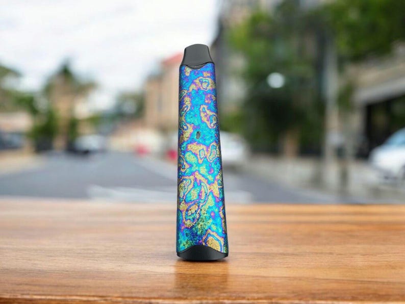 May include: A slender, iridescent vape pen with a black mouthpiece and base. The body of the pen features a swirling pattern of blues, greens, and yellows. The pen is standing upright on a wooden surface, with a blurred street scene in the background.