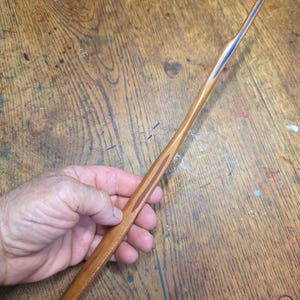 May include: A long, slender, tapered wooden wand with a smooth, polished finish. The wand features a light brown wood grain throughout, with a distinct dark brown inlay along the handle and a thin, vibrant blue accent line running near the tip.