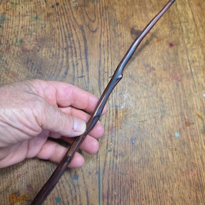 May include: A long, slender, dark brown wooden wand with a smooth, polished finish. The wand features a natural, slightly irregular branch-like shape with subtle knots and a tapered point at one end, showcasing a rich, deep wood grain texture.