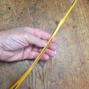 May include: A long, slender, hand-carved wooden wand with a smooth, polished yellow finish. The wand features a tapered design with a thicker center section and a pointed tip, accented by a thin, dark brown inlay stripe running along the handle.