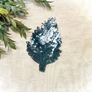 May include: A polished stone carving shaped like a serrated leaf, featuring a deep forest green base color with prominent white, cloud-like mineral inclusions throughout the surface, displaying a natural vein pattern and a smooth, glossy finish.