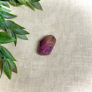 May include: A purple and brown gemstone with a unique swirling pattern. The stone is smooth and polished, and it has a natural, organic shape.