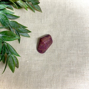 Könnte beinhalten: Ein tief purpurfarbener und rot gestreifter, getrommelter Stein mit einer glatten, polierten Oberfläche. Der Stein liegt auf einem beigefarbenen Stoffhintergrund.