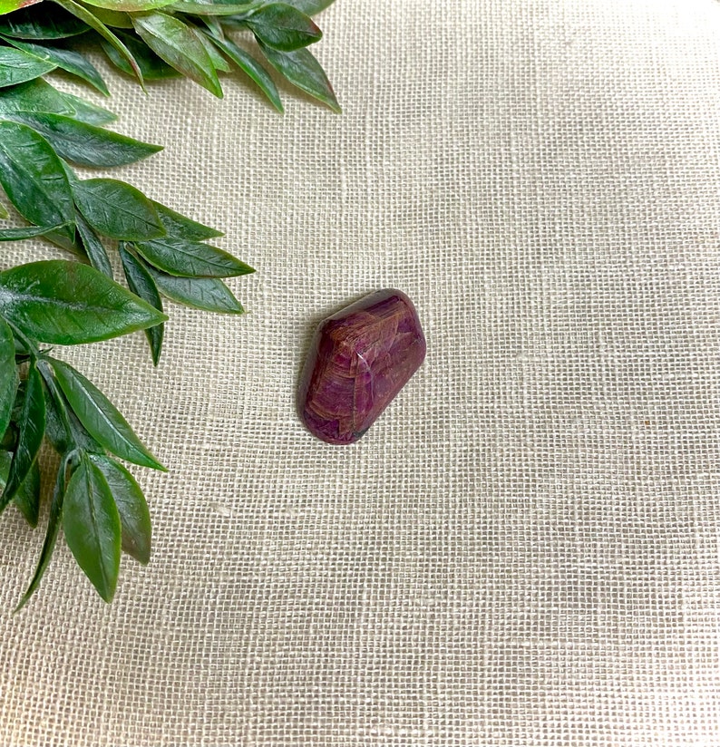 May include: A deep purple and red striped tumbled stone with a smooth, polished surface. The stone is resting on a beige fabric background.
