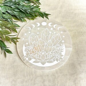 May include: A white selenite charging plate with a moon and crystal design. The plate features a crescent moon with three phases, a cluster of crystals, and a lotus flower.