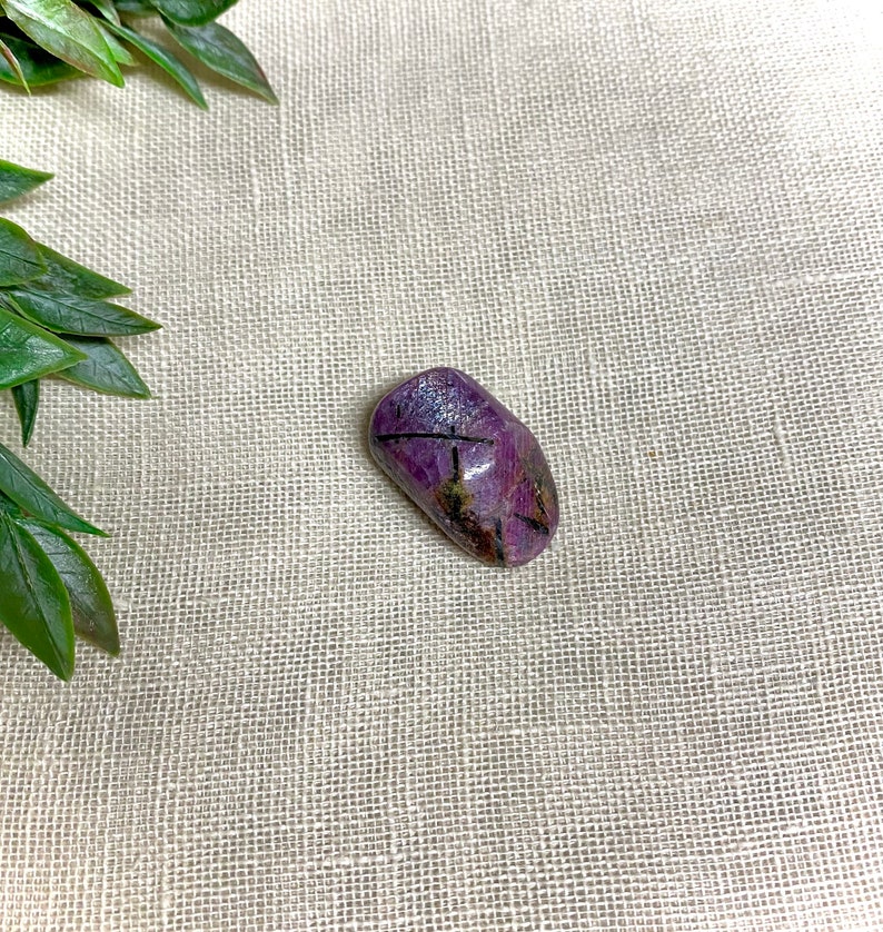 K&ouml;nnte beinhalten: Ein violetter und schwarzer Edelstein mit einer glatten, ovalen Form. Der Stein hat eine nat&uuml;rliche, polierte Oberfl&auml;che.