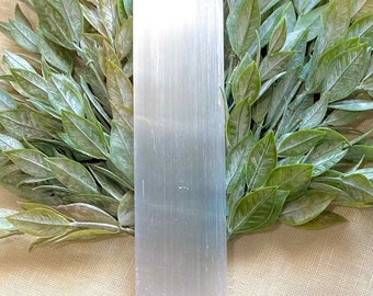 May include: A white selenite crystal wand, also known as a cleansing wand, is placed on a beige fabric background. The wand is surrounded by green leaves.