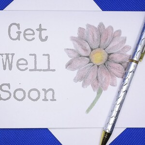May include: A white card with the words "Get Well Soon" printed in gray. A pink flower with a yellow center is drawn on the card. A silver pen with a gold top is lying on the card.