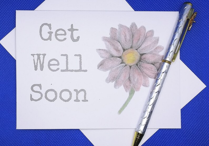May include: A white card with the words "Get Well Soon" printed in gray. A pink flower with a yellow center is drawn on the card. A silver pen with a gold top is lying on the card.