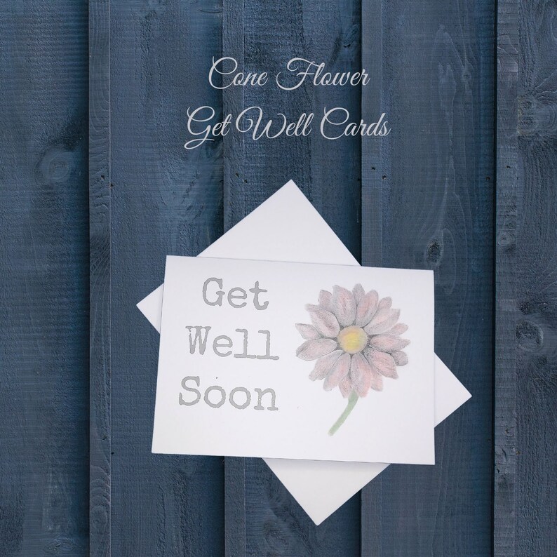 May include: A white card with the text "Get Well Soon" in gray lettering. A pink flower with a yellow center is drawn on the card. The card is on a blue wooden background.