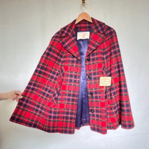 May include: A vintage red plaid cape coat with a navy blue lining and black buttons. The coat is hanging on a wooden hanger. A tag with the name "Saks" is attached to the coat.