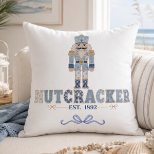 Könnte beinhalten: Ein weißes quadratisches Dekokissen mit einer blau-gold gemusterten Nussknacker-Grafik über dem Text NUTCRACKER EST. 1892 und einer blauen Schleifenverzierung. Das Design verwendet Schattierungen von Hellblau, Marineblau und Gold auf weißem Stoff.