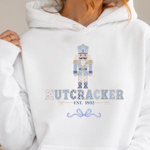 May include: A white hooded sweatshirt featuring a graphic of a nutcracker in shades of blue and white. Below the nutcracker, the text reads NUTCRACKER EST. 1892 in a blue and white patterned font, followed by a decorative blue ribbon graphic.