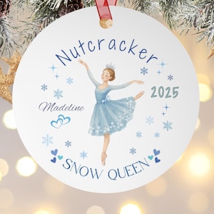 Op de afbeelding: Een witte, ronde kerstversiering met een ballerina-illustratie. De versiering heeft de woorden "Nutcracker" en "Snow Queen" in blauw schrift. De ballerina draagt een blauwe jurk en een kroon. Het jaar "2025" staat er ook op.
