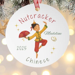 May include: White round Christmas ornament with a red ribbon. The ornament features a dancer in a yellow and red Chinese costume, the word "Nutcracker", the name "Madeline", the year "2025", and the word "Chinese".