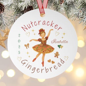 May include: A white Christmas ornament with a ballerina illustration. The ornament has the words "Nutcracker Gingerbread" and the year "2025" printed on it. The ballerina is wearing a brown dress and red shoes. A red ribbon is attached for hanging.