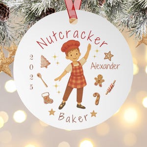 Könnte beinhalten: Ein weißer runder Weihnachtsschmuck mit rotem Band. Er zeigt einen Cartoon-Bäcker mit Kochmütze und Schürze, mit dem Text "Nutcracker Baker" und dem Namen "Alexander". Lebkuchenmänner, Sterne und eine Zuckerstange schmücken den Anhänger.