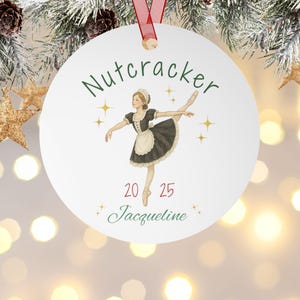 May include: A white Christmas ornament with a ballerina illustration. The ornament features the word "Nutcracker" in green script, the year "2025", and the name "Jacqueline". It is hanging from a red ribbon.