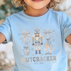 May include: A light blue t-shirt with a Nutcracker design, including bows, ballet shoes, and a crown. The text "NUTCRACKER EST. 1892" is printed below. The design is in shades of blue and silver.