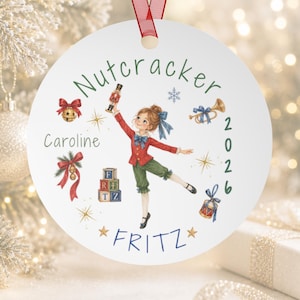 May include: A round white Christmas ornament featuring a central illustration of a child in a red jacket and green pants holding a nutcracker. The text reads Nutcracker, Caroline, 2026, and Fritz, surrounded by festive icons like bells, a horn, and blocks.