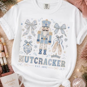 May include: White t-shirt with a blue and white Nutcracker design, including bows, ballet shoes, and the text "NUTCRACKER EST. 1892". The graphic features a detailed illustration of a Nutcracker soldier. Festive holiday clothing.