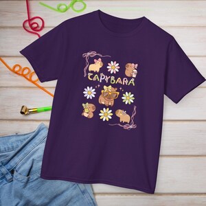 May include: A purple t-shirt featuring a whimsical design with the word "CAPYBARA" and illustrations of capybaras, flowers, and bows. The capybaras are depicted with sunglasses and holding coffee cups. The shirt is made of a soft material.