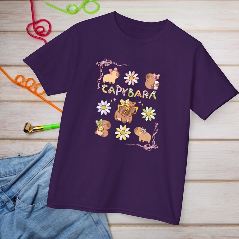 May include: A purple t-shirt featuring a whimsical design with the word "CAPYBARA" and illustrations of capybaras, flowers, and bows. The capybaras are depicted with sunglasses and holding coffee cups. The shirt is made of a soft material.