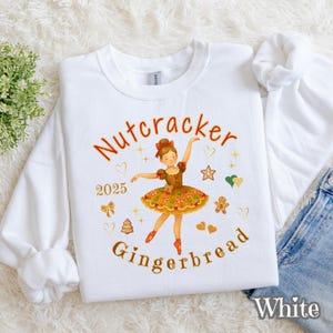 May include: White sweatshirt featuring a Nutcracker Gingerbread design. The graphic includes a ballerina in a brown and orange dress, the year 2025, and the words "Nutcracker Gingerbread" in orange script. Additional festive icons are scattered around the ballerina.