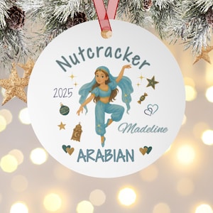 May include: Round white Christmas bauble with a red ribbon. It features a cartoon dancer in blue Arabian clothing, the word "Nutcracker", the year "2025", and the name "Madeline". Decorative stars and hearts surround the dancer.
