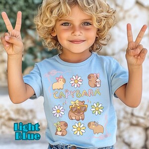 May include: Light blue t-shirt featuring a whimsical "Capybara" design with cartoon capybaras, flowers, and text. The shirt has short sleeves and is made of a soft material. The text "light blue" is at the bottom left of the shirt.