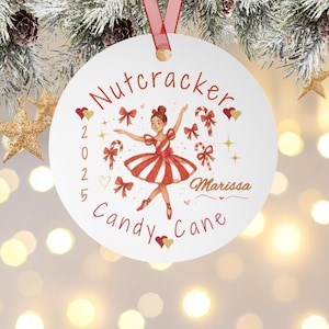 Puede incluir: Un adorno navideño blanco con una cinta roja. Presenta una bailarina con un vestido rojo y blanco, rodeada de bastones de caramelo y lazos. El texto dice "Nutcracker Candy Cane 2025 Marissa".