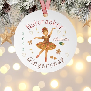 May include: A white Christmas ornament with a red ribbon, featuring a ballerina illustration. The ornament has the words "Nutcracker" and "Gingersnap" in red, along with the name "Isabella" and the year "2025".