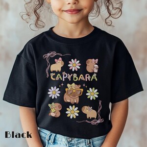 May include: Black t-shirt featuring a colorful graphic design with the word "CAPYBARA" and illustrations of capybaras, flowers, and bows. The shirt is made of a soft material and is suitable for children.